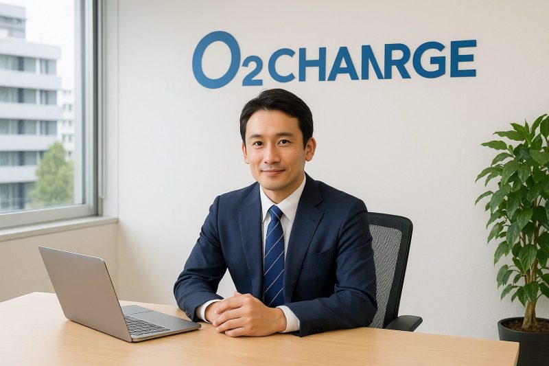 オーツーチャージ本社オフィスで日本人男性社員がスーツ姿でデスクに座り、ノートパソコンの前で穏やかに微笑む姿を16:9構図でリアルに表現した画像。信頼感と企業の専門性を感じさせるシーン